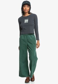 Dark gray long-sleeve crop top with graphic print, paired with green corduroy wide-leg pants, and brown lace-up shoes. Black cap worn.