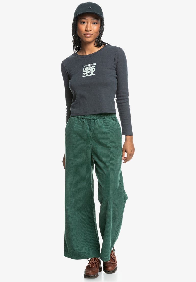Dark gray long-sleeve crop top with graphic print, paired with green corduroy wide-leg pants, and brown lace-up shoes. Black cap worn.