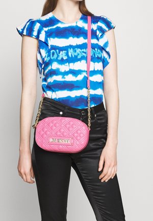 Prošívaná růžová crossbody taška se zlatým řetízkem, s logem na štítku, nošená s modrým tričkem s tie-dye vzorem a černými úzkými kalhotami.