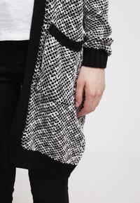 Černobílý pletený kardigan s texturovanou otevřenou vazbou, žebrovanými manžetami a detailem přední kapsy.