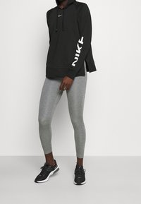 Czarna bluza z kapturem z białym napisem „NIKE” na rękawie, w połączeniu z jasnoszarymi legginsami i czarnymi butami sportowymi. Gładki materiał i dopasowany krój.