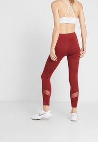 Donna che indossa un reggiseno sportivo bianco, leggings rossi a vita alta con pannelli in rete vicino ai polpacci e sneakers bianche Nike, in piedi su uno sfondo semplice.