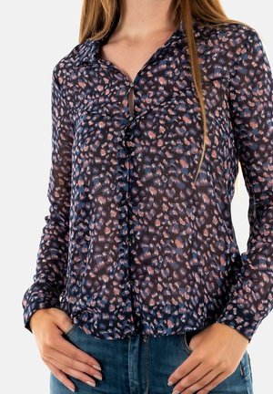 Femme portant un chemisier à manches longues, boutonné, de couleur sombre avec un motif abstrait bleu et rose, associé à un jean en denim bleu, les mains reposant près des poches.