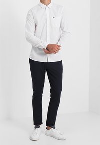 Camisa blanca de manga larga con botones y bolsillo en el pecho, combinada con pantalones oscuros y zapatillas blancas, posando frente a un fondo liso.