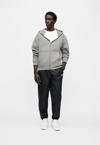 Γκρι φούτερ με φερμουάρ πάνω από λευκό μπλουζάκι, συνδυασμένο με μαύρο στενό παντελόνι και λευκά αθλητικά παπούτσια. Διαθέτει τσέπες και ελαστικά μανίκια.