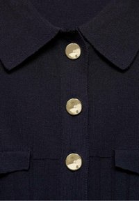 Chemise en maille bleu marine avec col, présentant trois boutons dorés brillants et une texture côtelée. Comprend deux poches sur le devant.