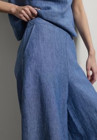 Persona que lleva puestos pantalones de lino azules, sueltos y de talle alto, y un top a juego, con una mano sujetando la tela cerca de la cintura.