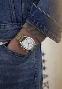 Montre chronographe avec un cadran blanc, un boîtier en argent et un bracelet en nylon vert. Portée sur un poignet avec une veste en denim et un pull beige.