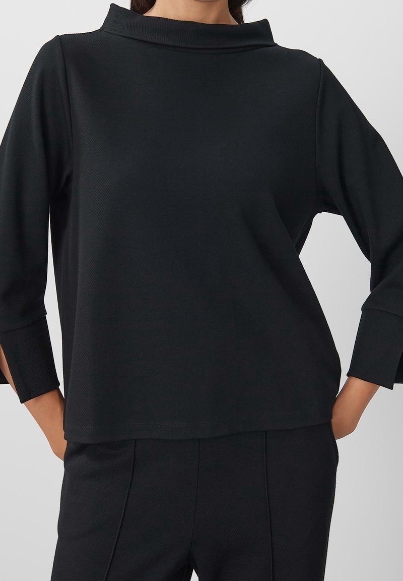 Pull-over noir texturé avec un col haut et des manches 3/4, présentant une coupe décontractée et un tissu lisse. Assorti à un pantalon foncé de même ton.