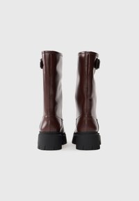 Botas marrones brillantes hasta la rodilla con una suela negra gruesa y un detalle de hebilla en la parte posterior, con una textura suave y un diseño aerodinámico.
