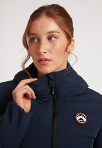 Giacca puffer blu navy con colletto alto, dotata di zip frontale e patch con marchio sul lato sinistro del petto. Materiale liscio e isolato.