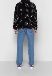 Personne portant une veste polaire noire avec des motifs de fissures blanches, un jean bleu droit et des baskets blanches avec des touches violettes, vue de dos.