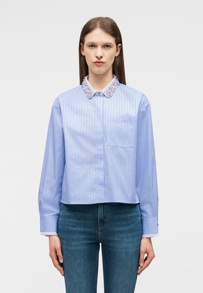 Γυναίκα που φοράει ανοιχτό μπλε, μακρυμάνικο, κοντό ριγέ πουκάμισο με διακοσμητικό γιακά και σκούρο μπλε τζιν, στέκεται μπροστά σε απλό φόντο.