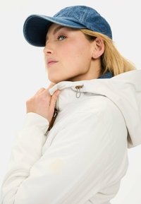 Witte jas met capuchon, voorzien van een ritssluiting en lichte beige accenten. Blauwe denim pet met een gebogen klep. Model staat poseert met handen bij de kraag.