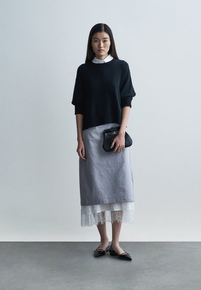 Zwarte gebreide trui, grijze midirok met kanten zoom, zwarte clutch en zwarte platte schoenen. Gladde texturen en gelaagd ontwerp.