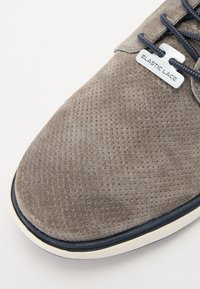 Grå semsket skinnsko med perforert tekstur, med marineblå elastiske lisser og en etikett merket "ELASTISKE LISSE." Rund tå, hvit mellomsole.