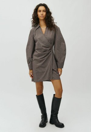Woman with curly hair wearing a brown plaid wrap dress and black knee-high boots standing against a plain light background.
