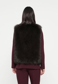 Gilet en fausse fourrure sombre avec un col haut, présentant une texture épaisse et douce. Porté par-dessus un haut bordeaux, avec une coupe droite et sans fermetures visibles.