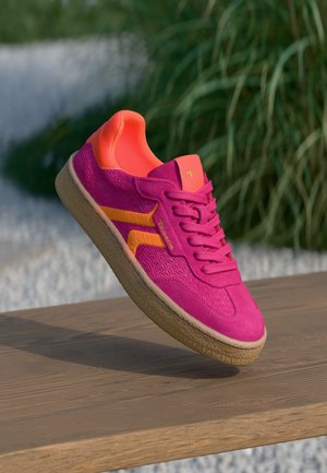 Lyserød sneaker med orange accenter og tekstureret sål, der svæver over en træoverflade udendørs med grønne planter i baggrunden.