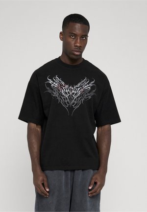 Homme portant un t-shirt noir oversize avec un motif abstrait de flamme argentée et rouge sur la poitrine, assorti à un pantalon ample gris foncé.
