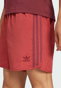 Pantaloni corti sportivi rossi Adidas con tre strisce rosso scuro sul lato e il logo vicino all'orlo, indossati con una maglietta rossa abbinata.