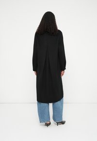 Mujer con cabello rizado vestida con un abrigo largo negro, jeans anchos azules y tacones negros de punta, de espaldas sobre un fondo blanco.