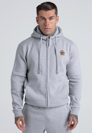 Mann trägt hellgrauen Kapuzenpullover mit Reißverschluss und passende Jogginghose, steht vor schlichtem hellem Hintergrund und blickt nach vorn.