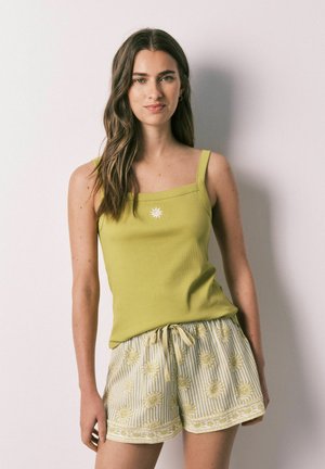 Giovane donna con capelli lunghi che indossa un top verde senza maniche e pantaloncini chiari con motivi solari, in piedi contro una parete chiara e liscia.