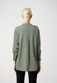 Camisa de manga longa verde-claro, texturizada, com punhos abotoados e detalhe de yoke nas costas, combinada com calças pretas.