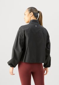 Gekürzte schwarze Jacke mit hohem Kragen, elastischen Bündchen und einem dezenten Logo-Detail auf dem Rücken, mit glatter Textur und leichtem Material.