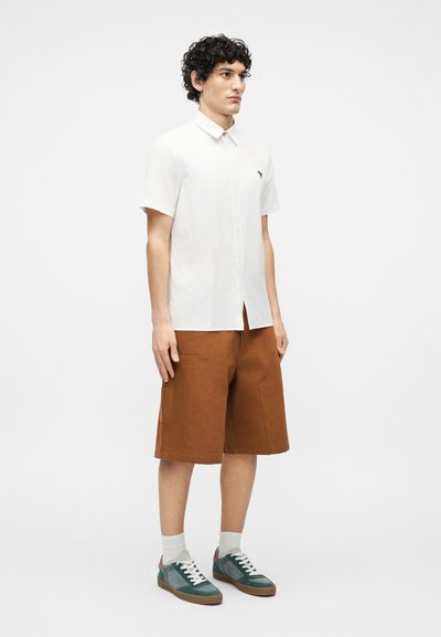 Man standing wearing a white short-sleeve button-up shirt, brown knee-length shorts, white socks, and teal sneakers on a white background.