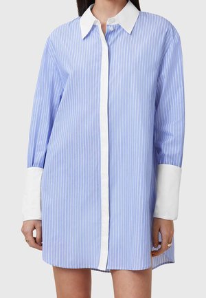 Woman wearing a long blue shirt dress with white vertical stripes, a white collar, and white wide cuffs, standing against a plain background.