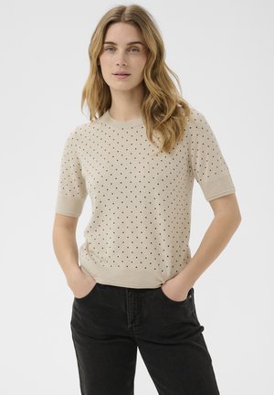 Femme aux cheveux châtain clair ondulés porte un pull beige à manches courtes avec des pois noirs et un pantalon noir, mains dans les poches, fond neutre.