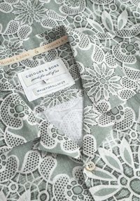 Camicia floreale verde con motivo in pizzo bianco, dotata di colletto, etichetta con la scritta "COLOURS & SONS" e un bottone al collo.