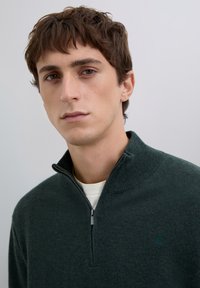 Maglione verde scuro con zip e colletto alto, realizzato in tessuto testurizzato. Presenta un logo discreto sul petto e uno strato interno bianco a contrasto.