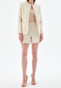 Tkaninowy żakiet w kratę z okrągłym dekoltem i dwoma przednimi kieszeniami, w połączeniu z pasującymi spodniami w paski. Kolor jasnobeżowy, gładka tekstura.
