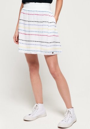 White skirt with horizontal rows of small multicolored dots, model standing with hands partially in pockets, wearing white high-top sneakers.