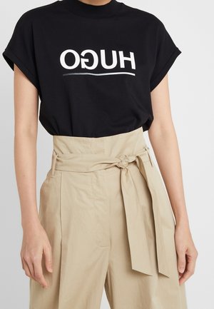 Mustaa puuvillapooloa, jossa on lyhyet hihat ja valkoinen HUGO-teksti. Yhdistetty korkeavyötäröisiin beigenvärisiin housuihin, joissa on solmittava vyö.