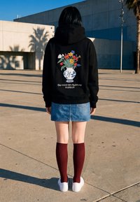 Personne portant un sweat à capuche noir avec un imprimé vase à fleurs coloré et un texte, jupe en jean, chaussettes hautes bordeaux et baskets blanches, debout à l'extérieur.