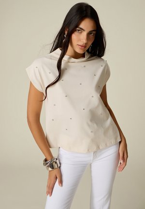 Femme aux longs cheveux foncés portant un haut beige clair avec de petites décorations et un pantalon blanc, accessoirisée avec des bracelets argentés au poignet gauche.