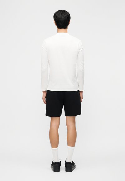 Άνδρας που στέκεται με την πλάτη, φοράει λευκό πουκάμισο μακρυμάνικο, μαύρο σορτς, λευκές κάλτσες και μαύρα αθλητικά παπούτσια σε απλό φόντο.