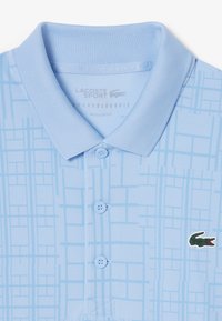 Polo shirt bleu clair avec un motif de grille texturé, col traditionnel et trois boutons. Présente un logo crocodile vert sur la poitrine.