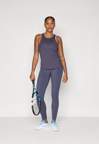 Sportos öltözék, amelyben egy formázott sötétlila trikó és az ahhoz illő leggings található. Kék részletekkel ellátott teniszütőt tart a kezében. Fehér sportcipők.