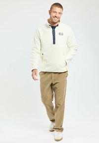 Pull en polaire blanc avec col et boutons navy, poches zippées avec accents jaunes, associé à un pantalon en velours côtelé marron clair et des baskets blanches.