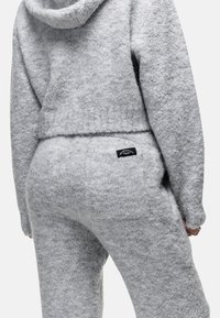Zachte, grijze, gestructureerde gebreide hoodie en bijpassende broek met een zwart rechthoekig label op de achterzak, gedragen door een persoon die van achteren is afgebeeld.