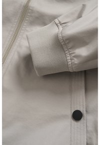 Hellbeige Jackenstoff mit glatter Textur. Verfügt über einen Reißverschluss und gerippte Bündchen. Enthält ein Detail mit schwarzem Druckknopf.