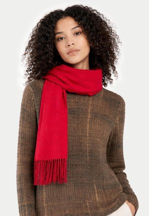 Frau mit lockigem Haar, die einen braunen, strukturierten Pullover und einen leuchtend roten, mit Fransen versehenen Schal um den Hals trägt.