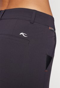 As calças de tecido escuro apresentam um corte slim, costura visível, um bolso traseiro e um pequeno logotipo branco com um destaque laranja perto da cintura.