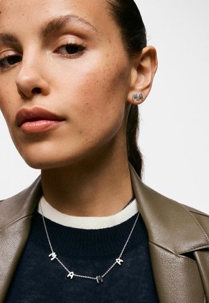 Primer plano de una mujer que lleva pendientes plateados con las letras "MAMA" y un collar a juego con las letras "MAMA", con una camiseta en blanco y negro y una chaqueta de cuero marrón.