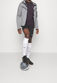 Chaqueta gris con cremallera, camiseta negra con patrón morado, pantalones cortos negros, calcetines blancos, botas negras, de pie sobre un balón de fútbol.